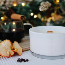 Load image into Gallery viewer, White candle in a glass holder with dried fruit and a glass of mulled wine on a table with a blurred Christmas tree in the background.