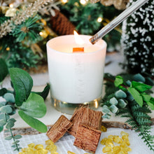Load image into Gallery viewer, Candle with a burning wick, surrounded by greenery and decorative elements on a wooden surface.