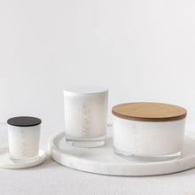 Load image into Gallery viewer, An image of a large candle with a wooden coloured lid, a medium candle with a white coloured lid and a small candle with a black coloured lid.  All three candles are in white glass vessels with MJM home fragrance logos