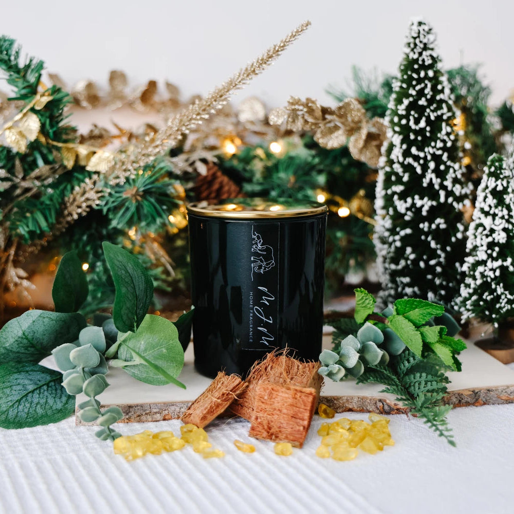 Green candle with a gold lid & MJM Home Fragrance branding.  Surrounded by ingredients that depict the scent notes such as wood, eucalyptus, pine and resin. All sitting in front of a festive background