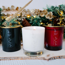 Load image into Gallery viewer, Three candles in emerald green, white, and red containers on a wooden board with Christmas decorations in the background.
