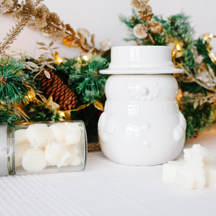 White ceramic snowman shaped wax melt burner with top hat lid on a white surface with Christmas decorations in the background