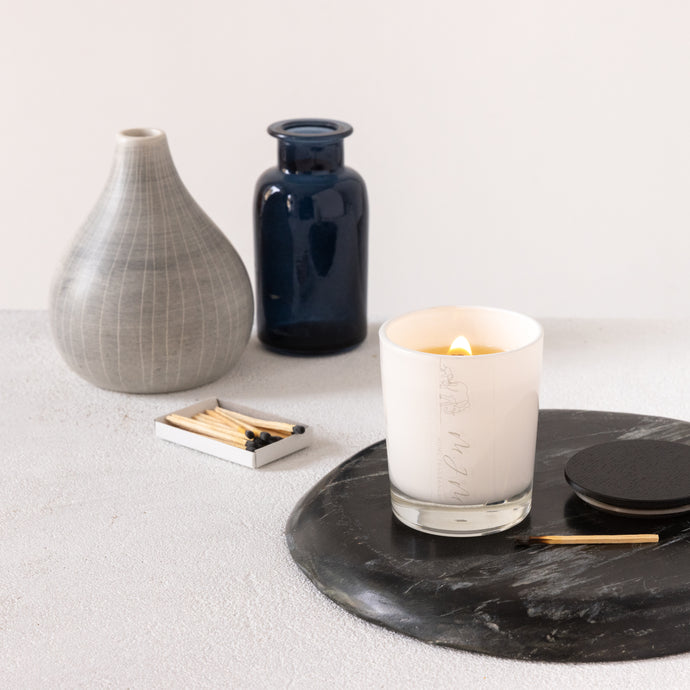 An image of a lit small candle with a wooden wick, inside a white glass vessel with MJM Home Fragrance logo